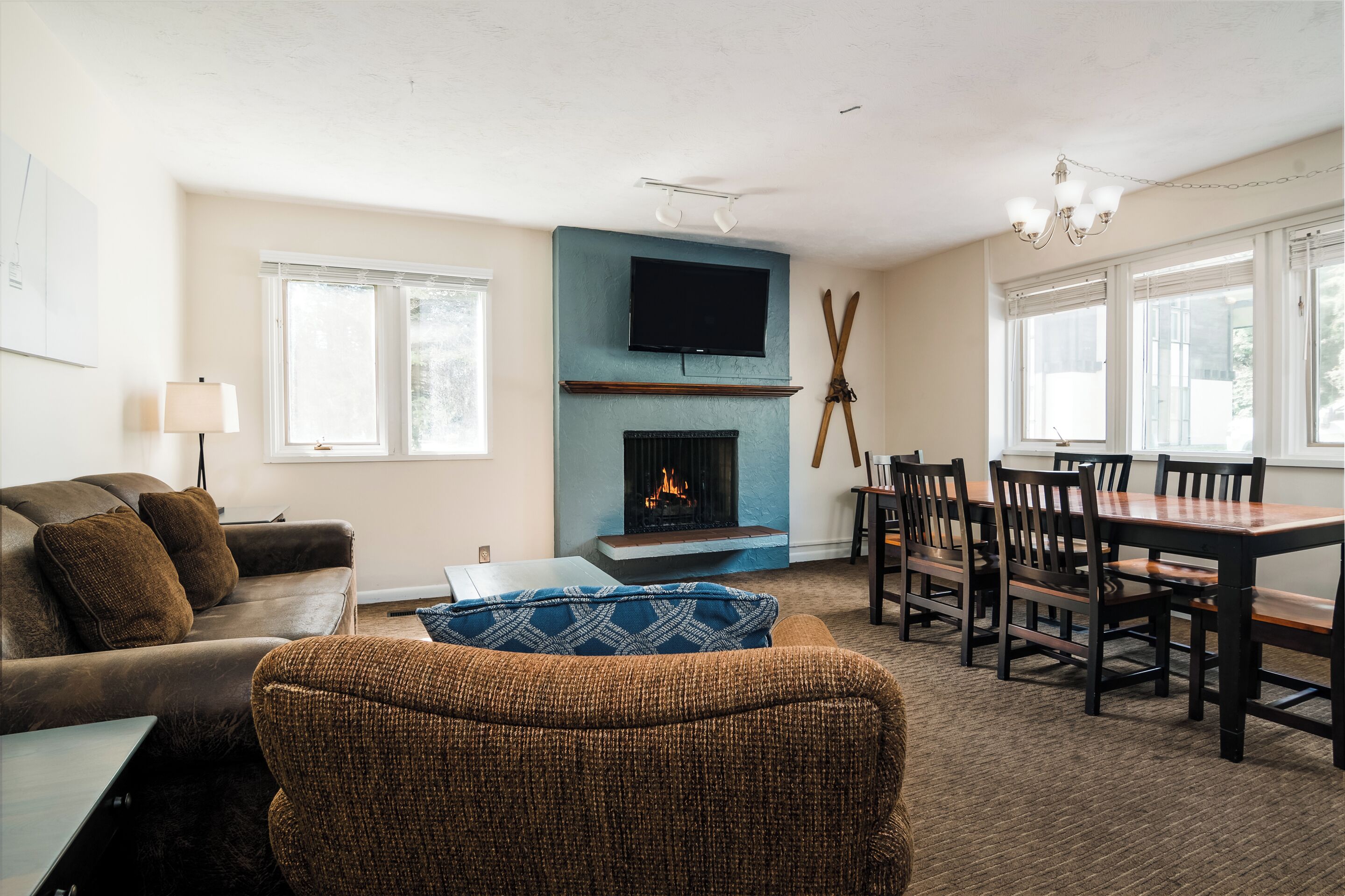 A living room with a fireplace, a television mounted above it, a dining area with a table and chairs, and a sofa with cushions.