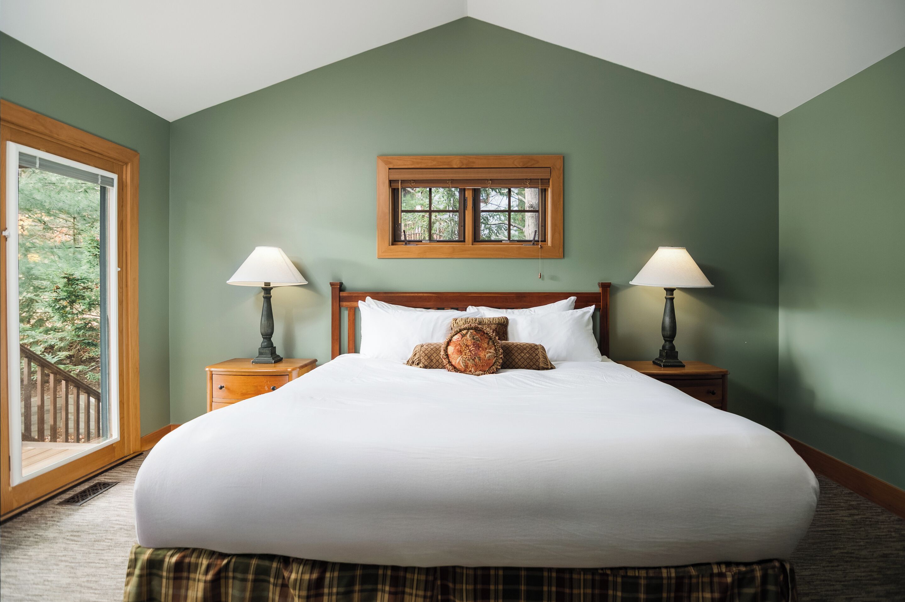 A large bed with white linens, two bedside lamps, a window with a wooden frame, and a view of a wooden deck.