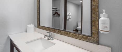 Une salle de bain avec un lavabo blanc, un miroir avec un cadre doré et un flacon de lotion pour le corps posé sur le mur.