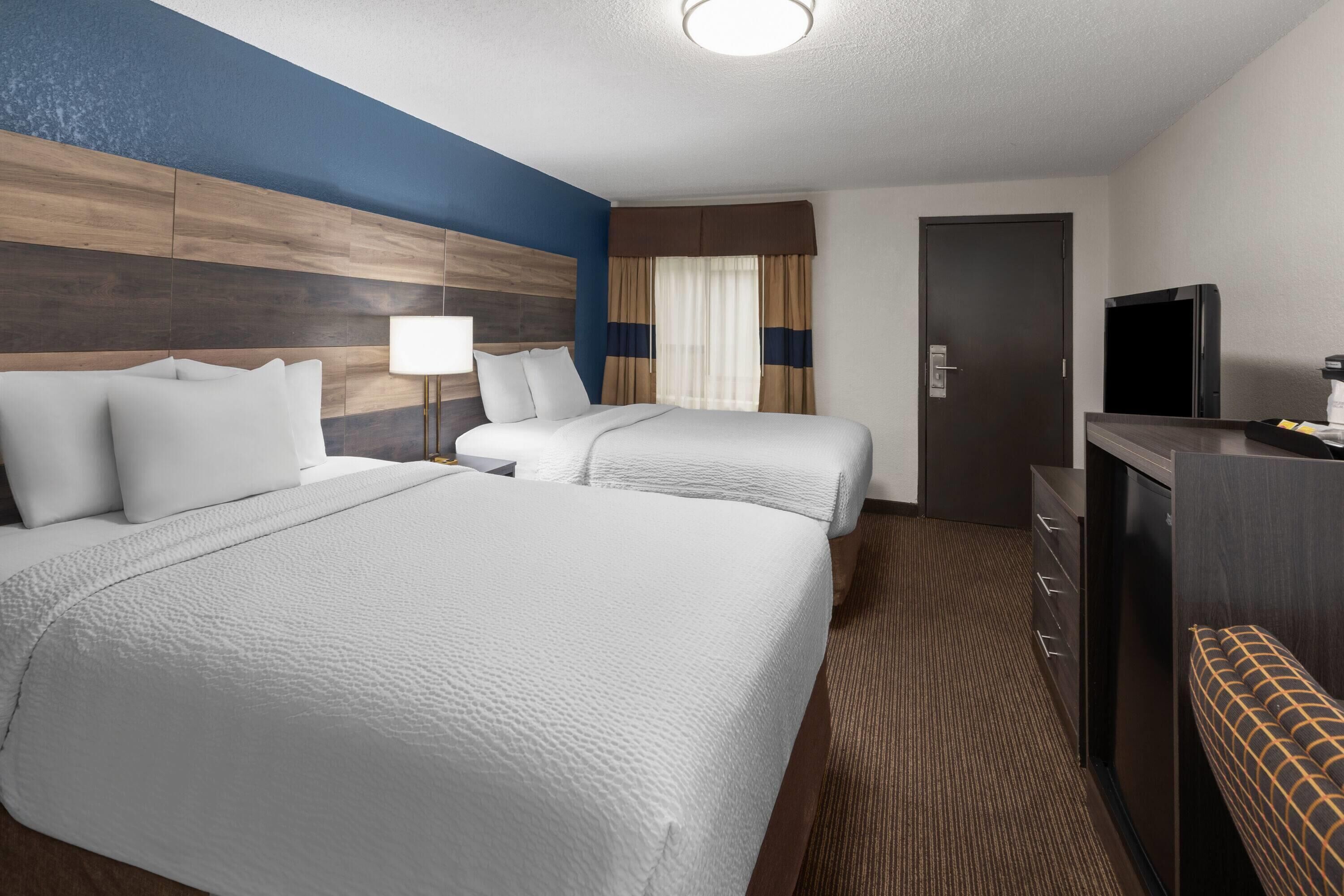 A hotel room with two beds, a desk, a television, and a bench.
