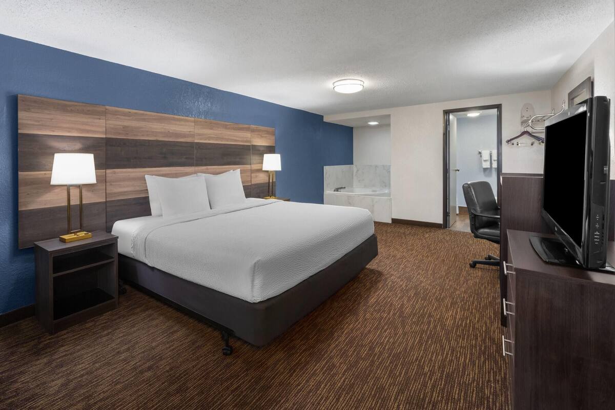 A hotel room with a large bed, two bedside tables with lamps, a television, a desk, and a bathroom visible through an open door.