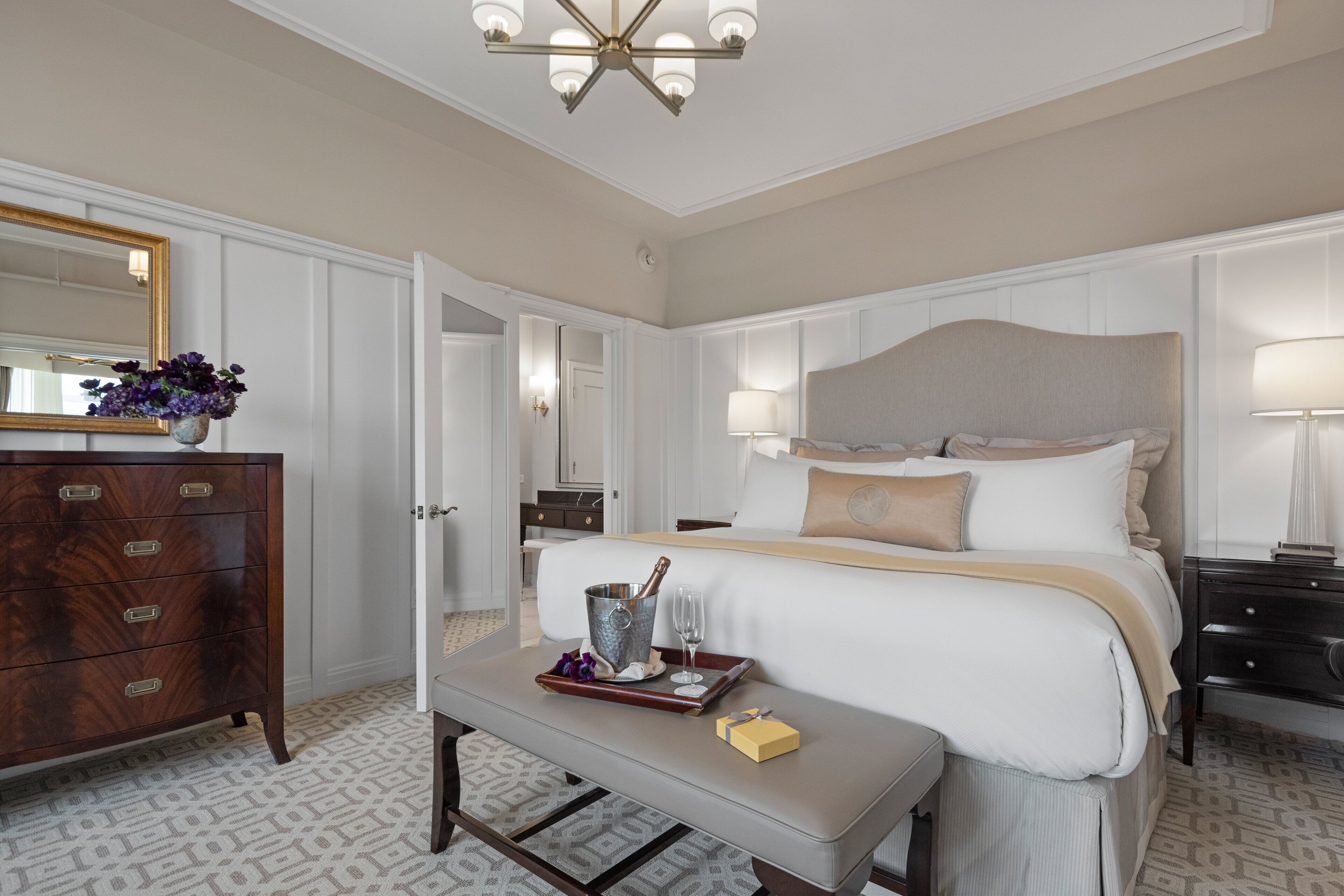 A hotel room with a bed, bedside tables, a dresser, a mirror, and a chandelier.