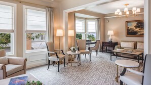 Uma sala de estar bem iluminada com um sofá, cadeiras, uma mesa de centro e um espaço de refeições. Há janelas amplas com cortinas, um candelabro e um quadro na parede.