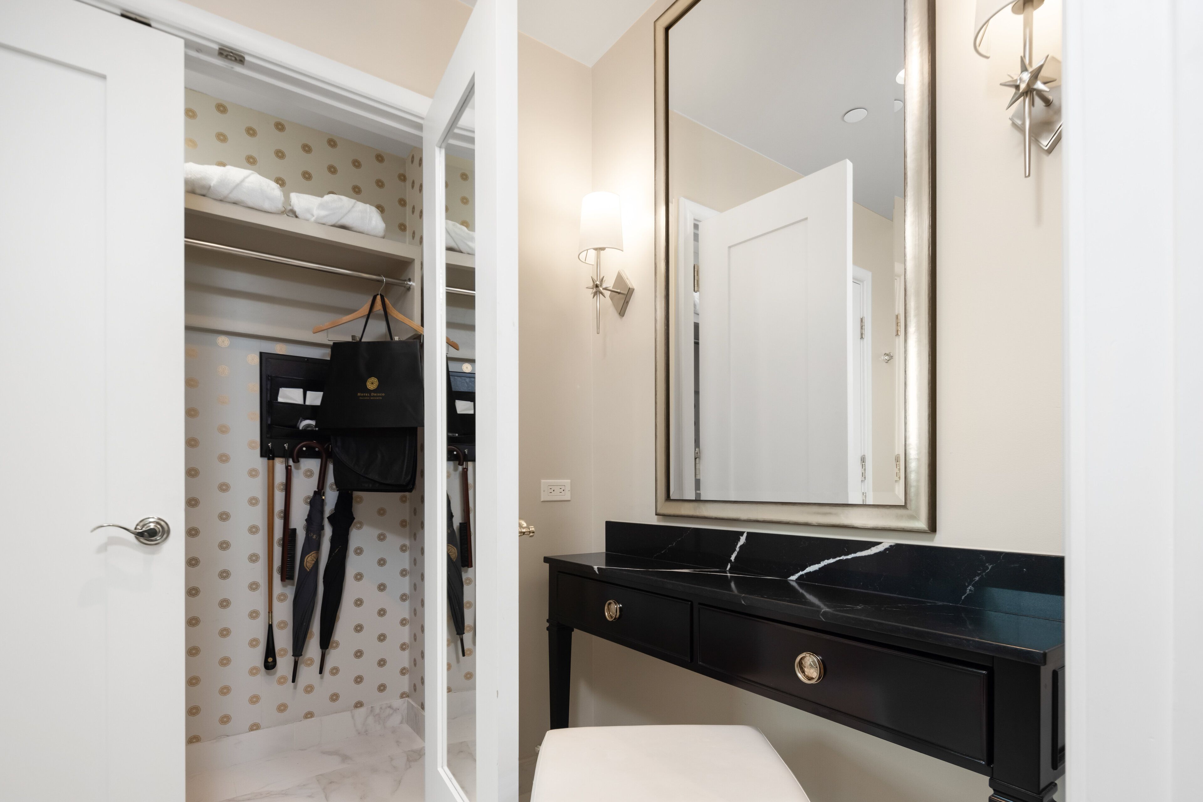 A hotel bathroom with a vanity, a mirror, and a walk-in closet with hanging items.