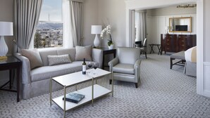 Quarto de hotel moderno com sofá, poltrona e mesa de centro. Oferece vista da cidade pela janela, um candelabro e um espelho na parede.
