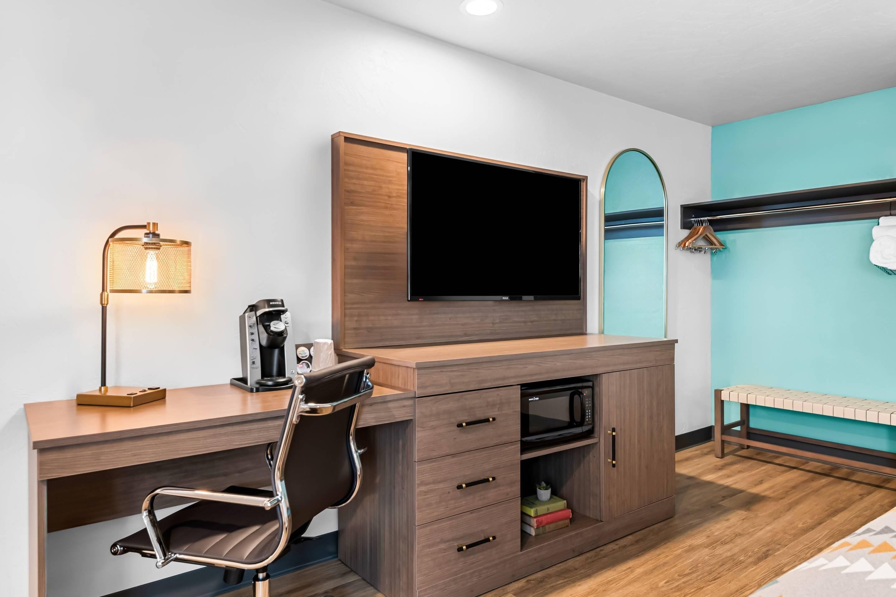A modern hotel room with a wooden desk, a flat-screen TV, a coffee maker, and a bench by the window.