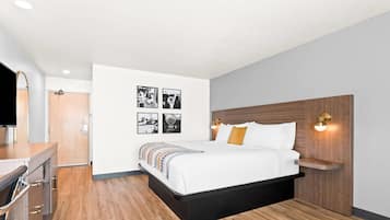 Una habitación de hotel moderna con piso de madera, cama con ropa blanca y almohadas naranjas, escritorio con televisor de pantalla plana y obras de arte colgadas en la pared.