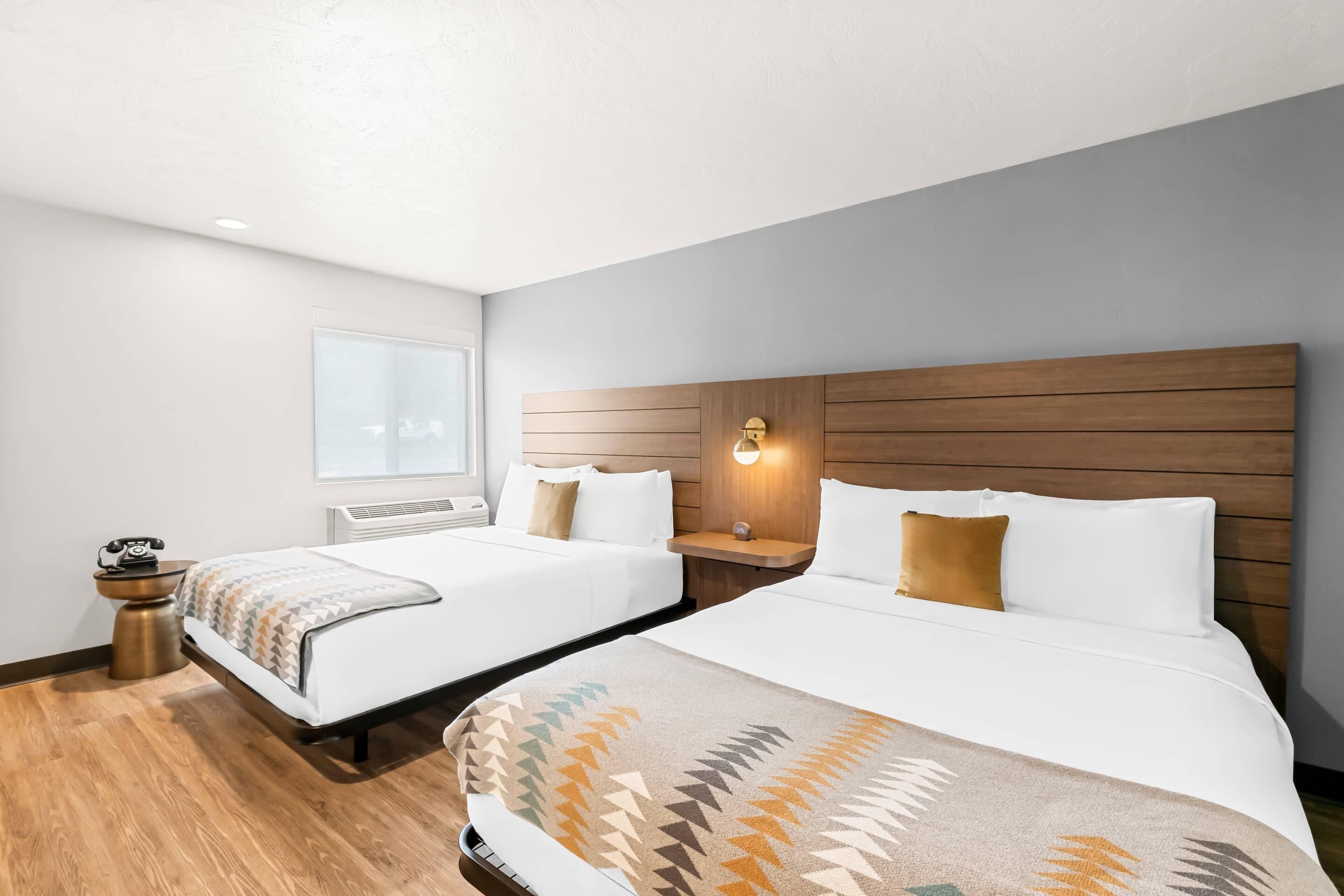 A hotel room with two beds, wooden headboards, a bedside table with a lamp, a small round table with a telephone, and a window with blinds.