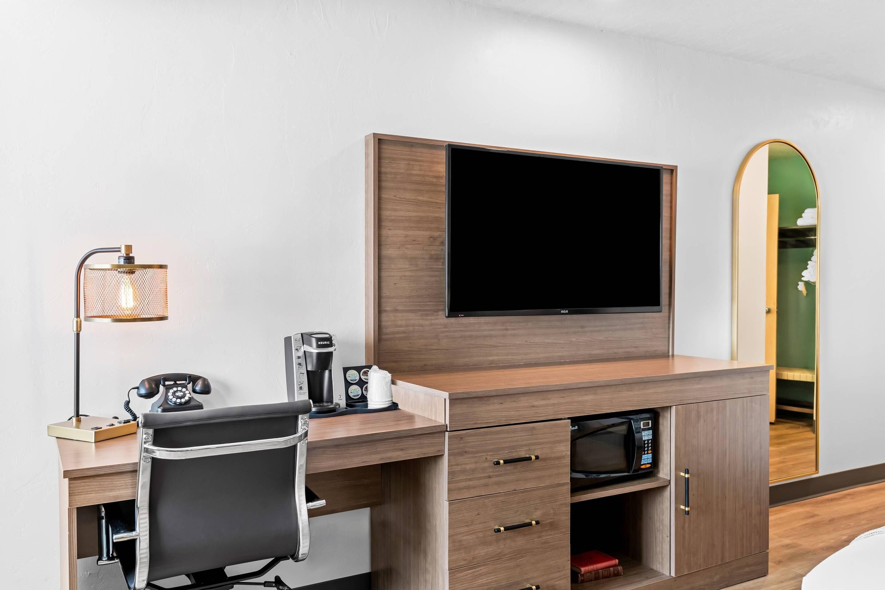 Ein Hotelzimmer mit Holztisch, Flachbildfernseher, Kaffeemaschine, Telefon und Spiegel.