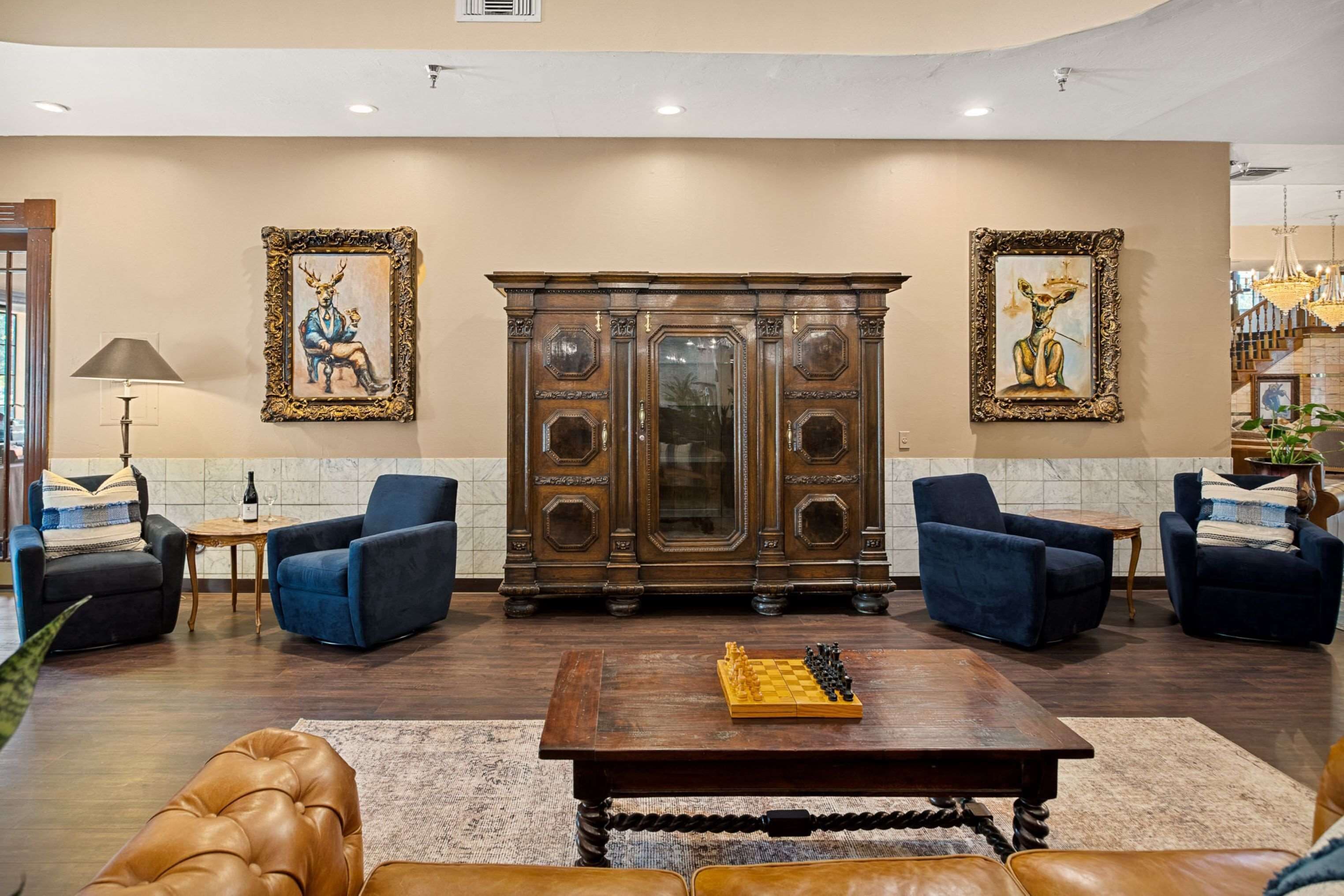 A spacious lobby with a large wooden cabinet, two blue armchairs, a leather sofa, and a chess table.
