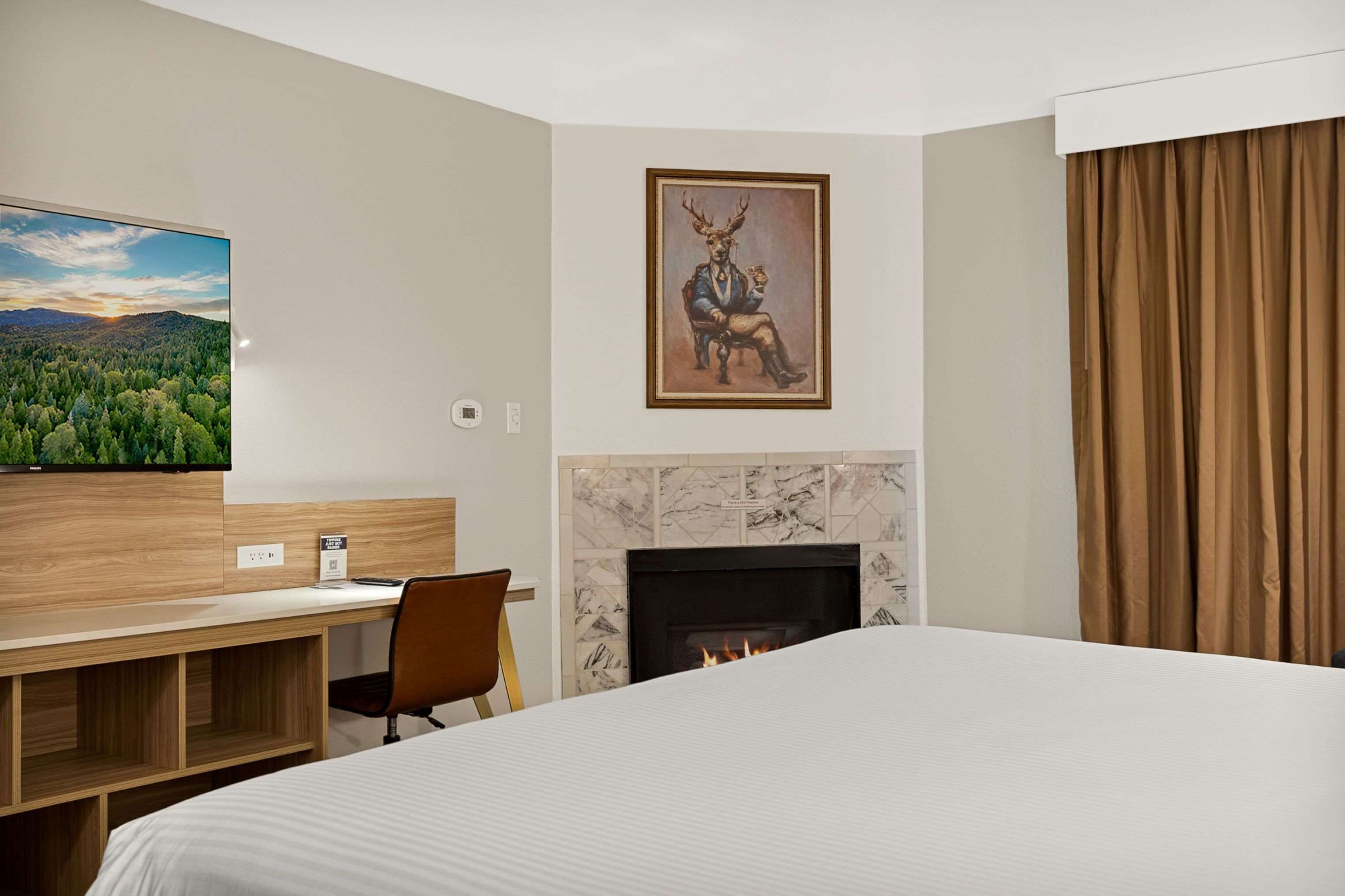 Habitación de hotel con cama, escritorio con silla, chimenea, un cuadro y vista a un bosque.