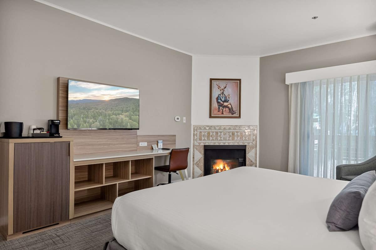 Une chambre d’hôtel dotée d’un grand lit, d’un bureau avec une chaise, d’un téléviseur à écran plat fixé au mur, d’une cheminée et d’une grande fenêtre avec des rideaux.