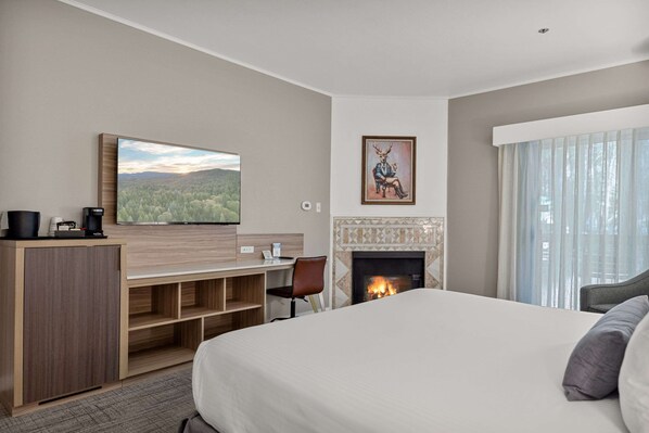Habitación de hotel con una cama grande, un escritorio con silla, un televisor de pantalla plana en la pared, chimenea y un ventanal con cortinas.