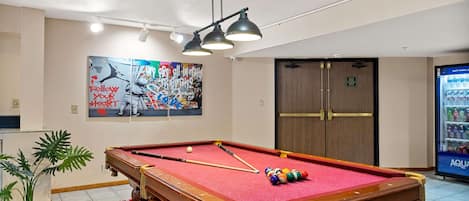A pool table with a red felt surface, surrounded by a red rug, in a room with a ceiling light fixture and a wall with a colorful poster.