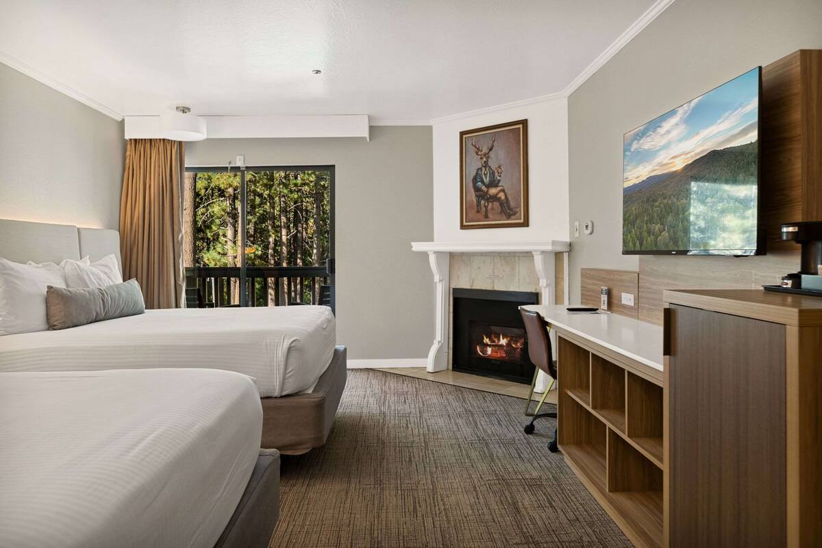 Une chambre d’hôtel avec deux lits, une cheminée, un bureau, une télévision et une grande fenêtre donnant sur la forêt.