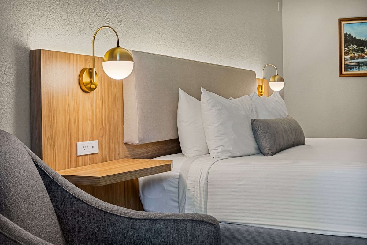 Une chambre d’hôtel avec une tête de lit en bois, une table de chevet, un fauteuil gris et un lit avec une literie et des oreillers blancs.