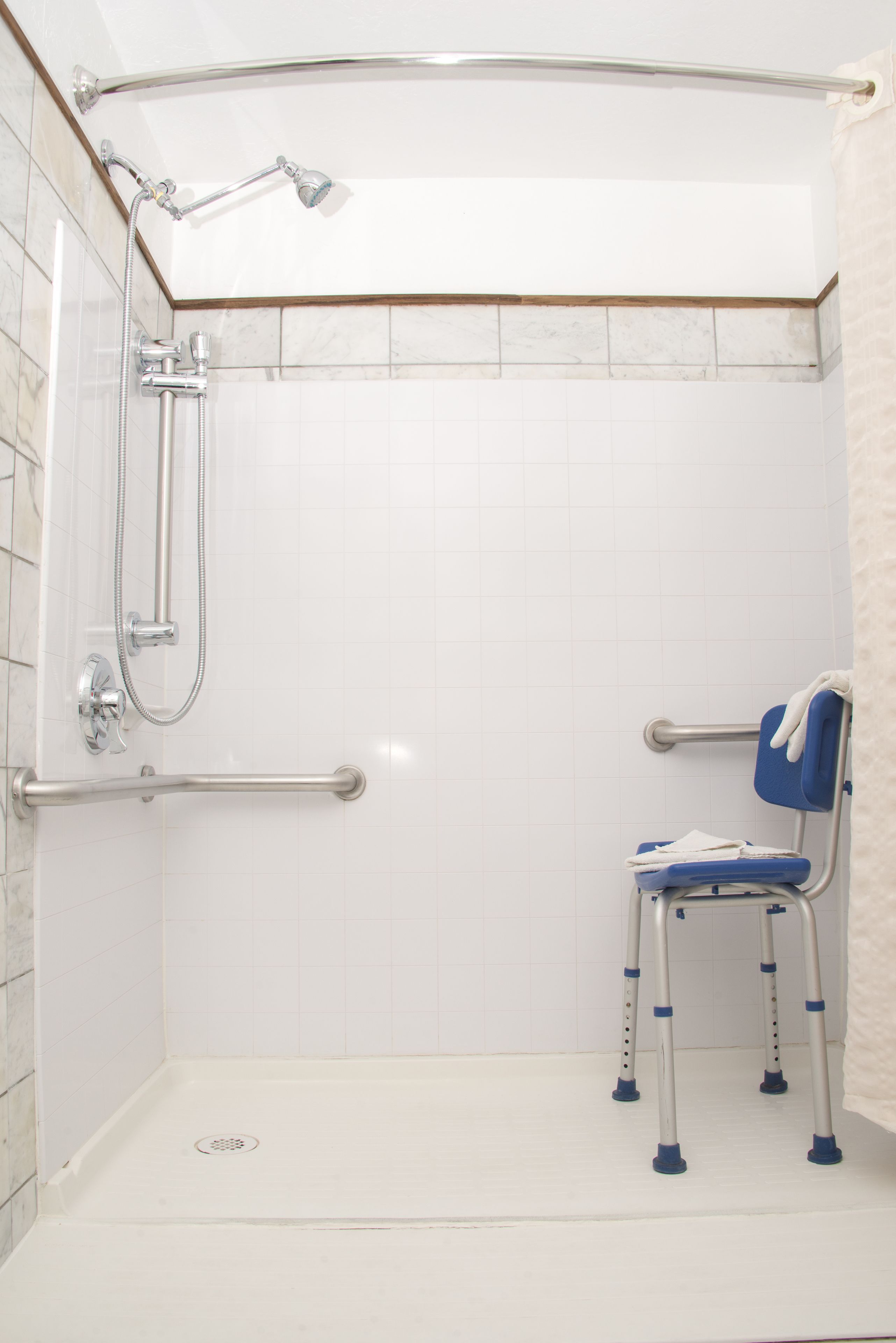 Une salle de bain avec des murs carrelés de blanc, une douche équipée d’un pommeau de douche à main, d’une barre d’appui et d’un banc accessible aux personnes en fauteuil roulant.