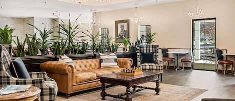 A spacious living room with a leather sofa, a chess table, and multiple chairs. There are several potted plants and a chandelier.