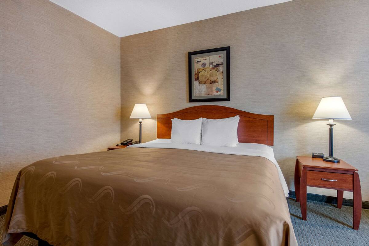 A hotel room with a large bed, two bedside tables with lamps, a wooden headboard, and a framed picture on the wall.
