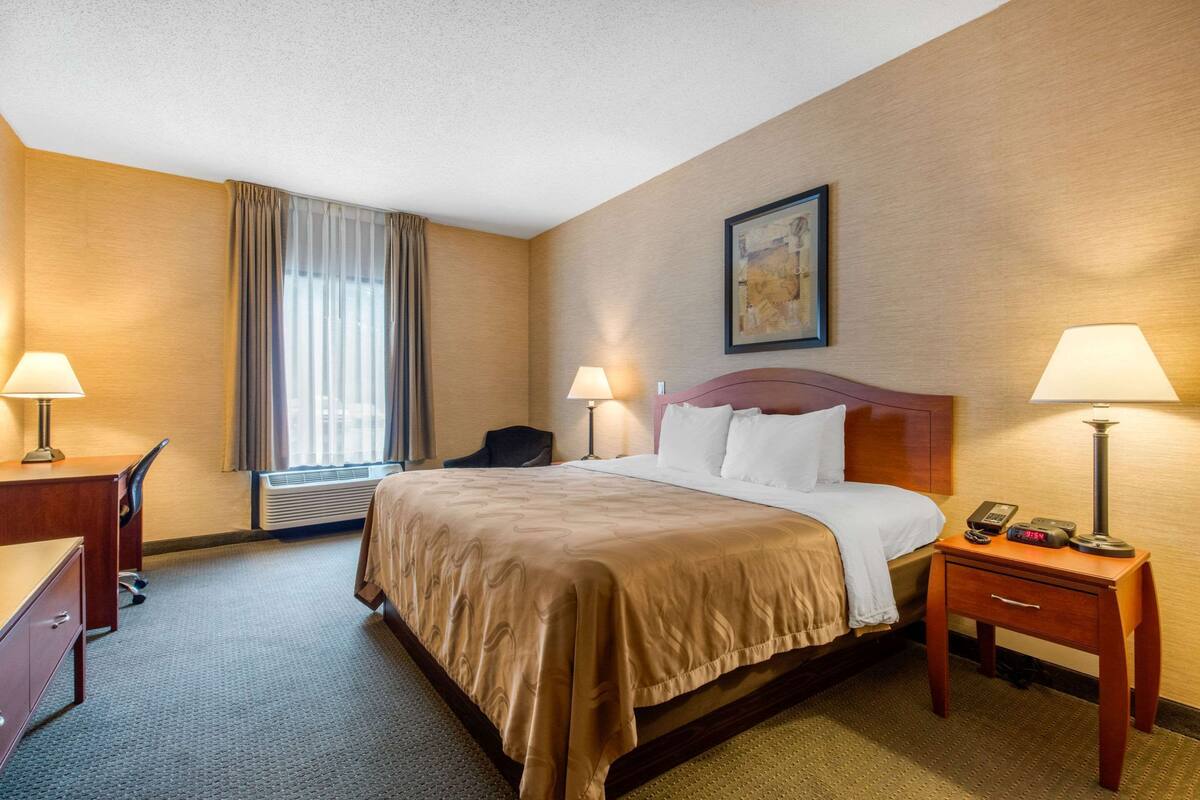 A hotel room with a bed, two bedside tables with lamps, a desk, a chair, and an air conditioning unit.