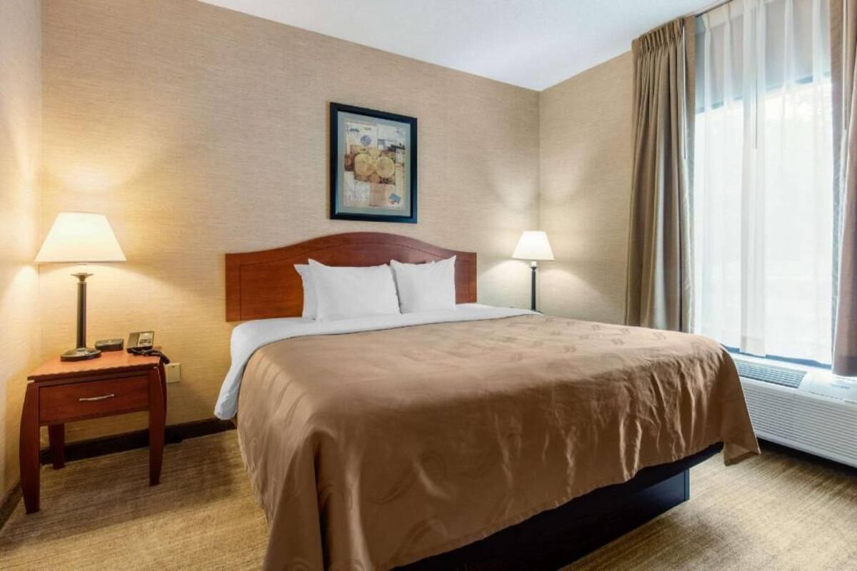 A hotel room with a bed, two bedside lamps, a nightstand, a framed picture on the wall, and a window with curtains.