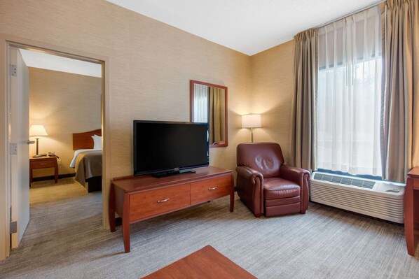 A hotel room with a flat-screen TV on a wooden console, a leather armchair, a large window with curtains, and a mirror on the wall. - Quality Inn & Suites Benton - Draffenville (Benton)