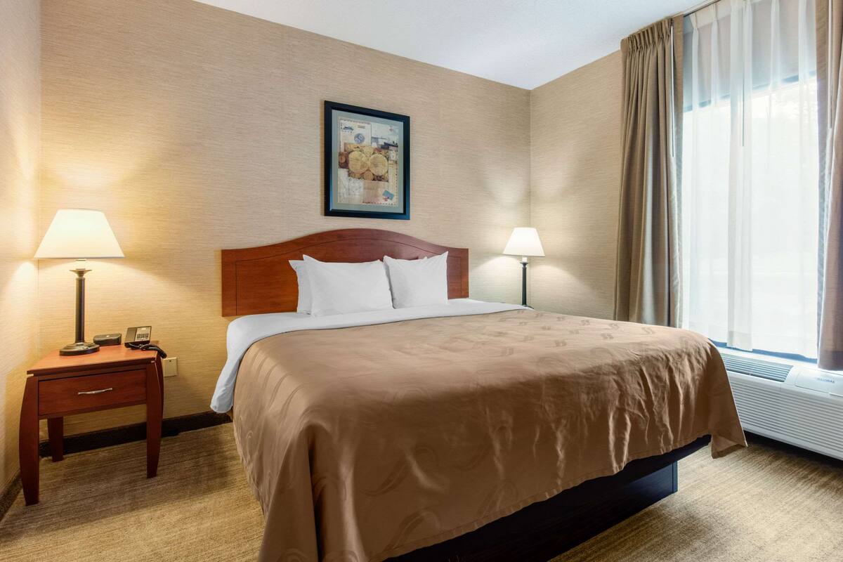 A hotel room with a bed, two bedside lamps, a nightstand, a framed picture on the wall, and a window with curtains.
