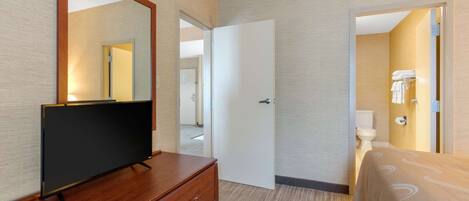 A hotel room with a wooden dresser, a flat-screen TV, a bed with a patterned bedspread, and a bathroom visible through an open door.