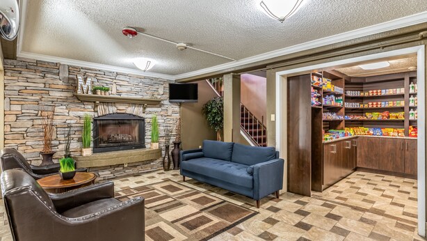 A living room with a stone fireplace, blue sofa, and a kitchen area with shelves stocked with snacks. - Park Inn by Radisson, Detroit Metro Airport (Romulus)