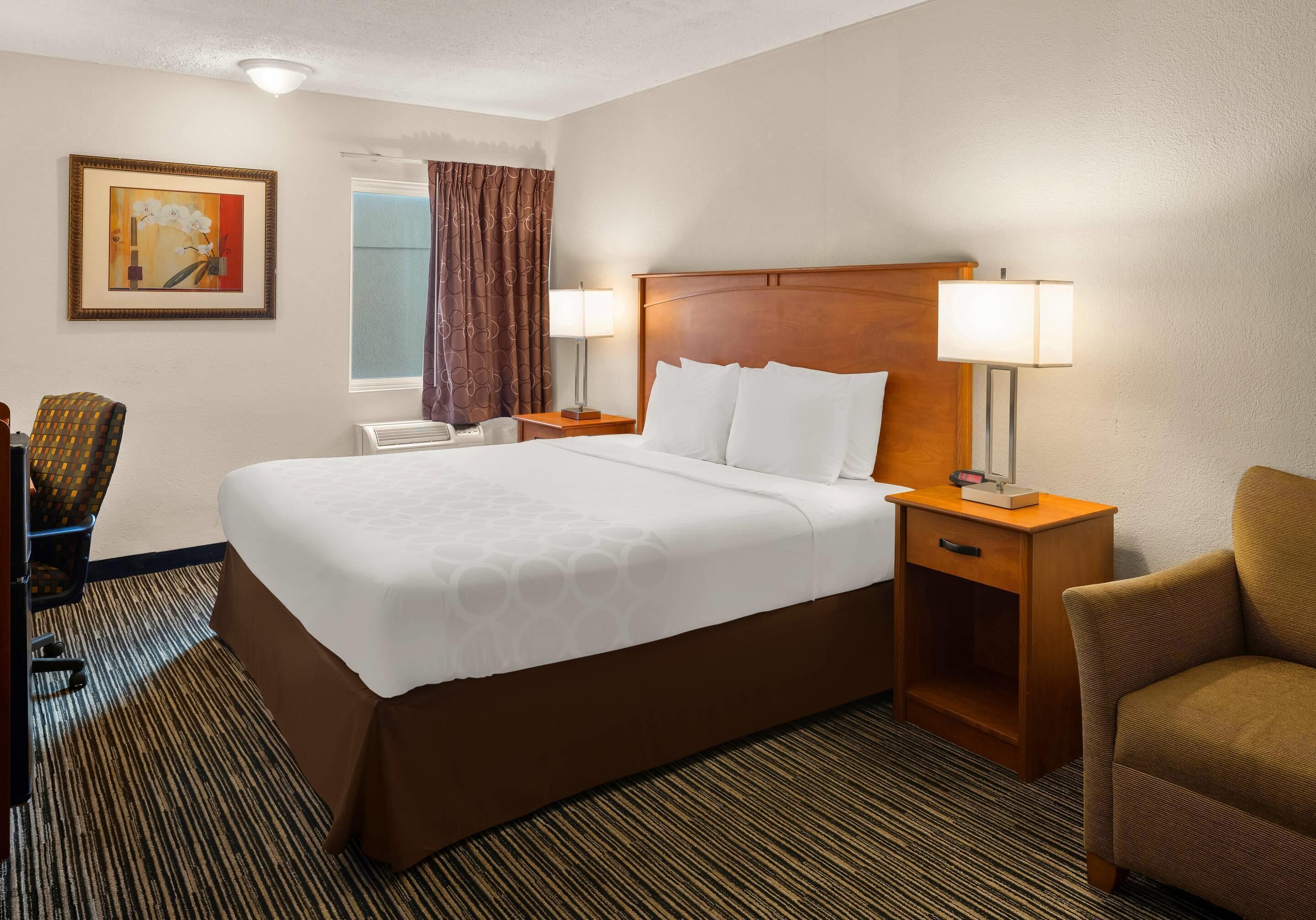 A hotel room with a large bed, two bedside tables with lamps, a chair, a desk, a window with curtains, and a painting on the wall.