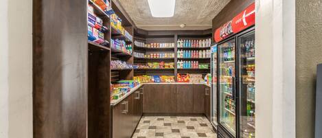 A narrow corridor with shelves stocked with snacks and beverages, a Coca-Cola refrigerator, and a tiled floor.