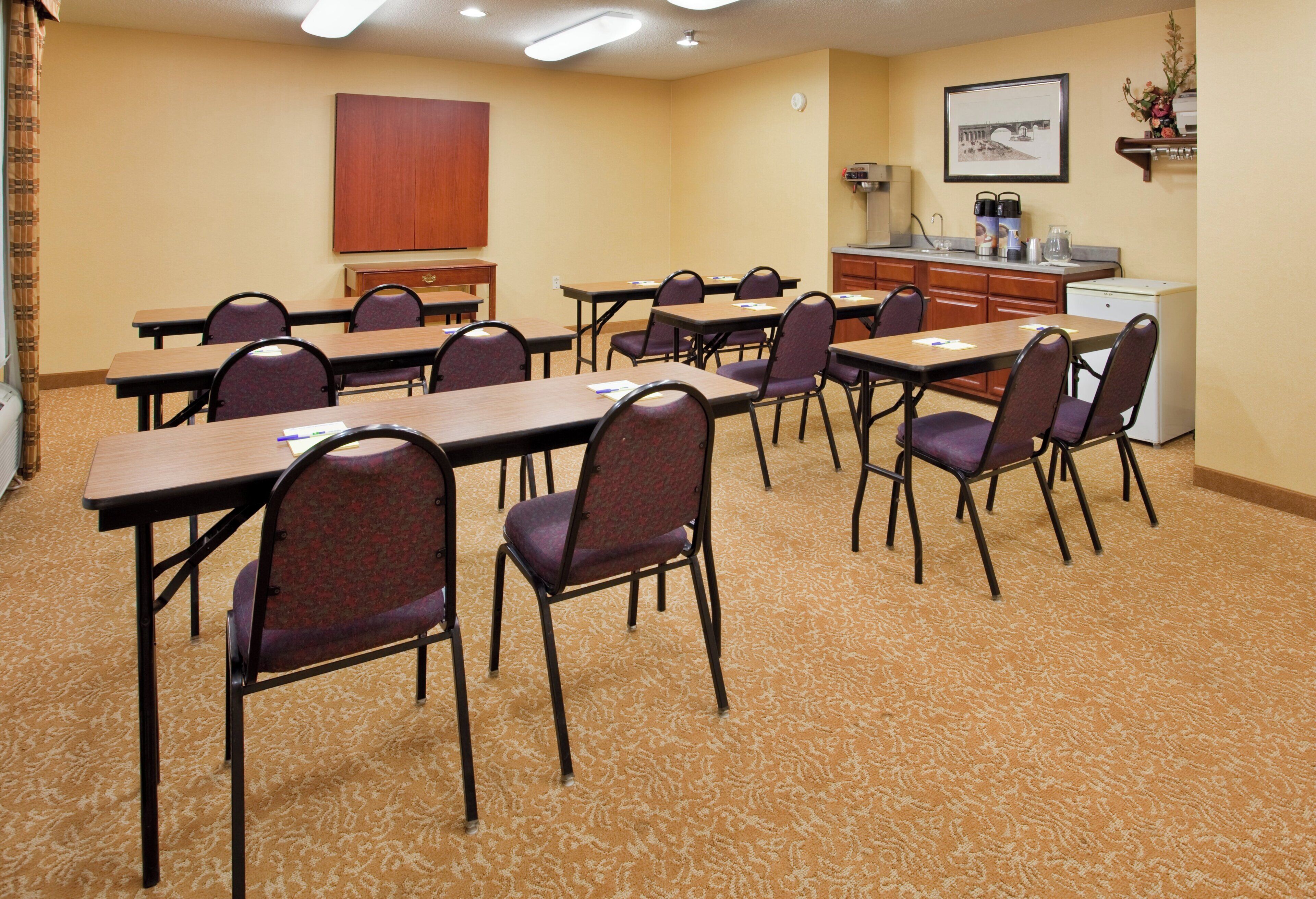 Un salón de conferencias con mesas y sillas, una kitchenette y un cuadro en la pared.