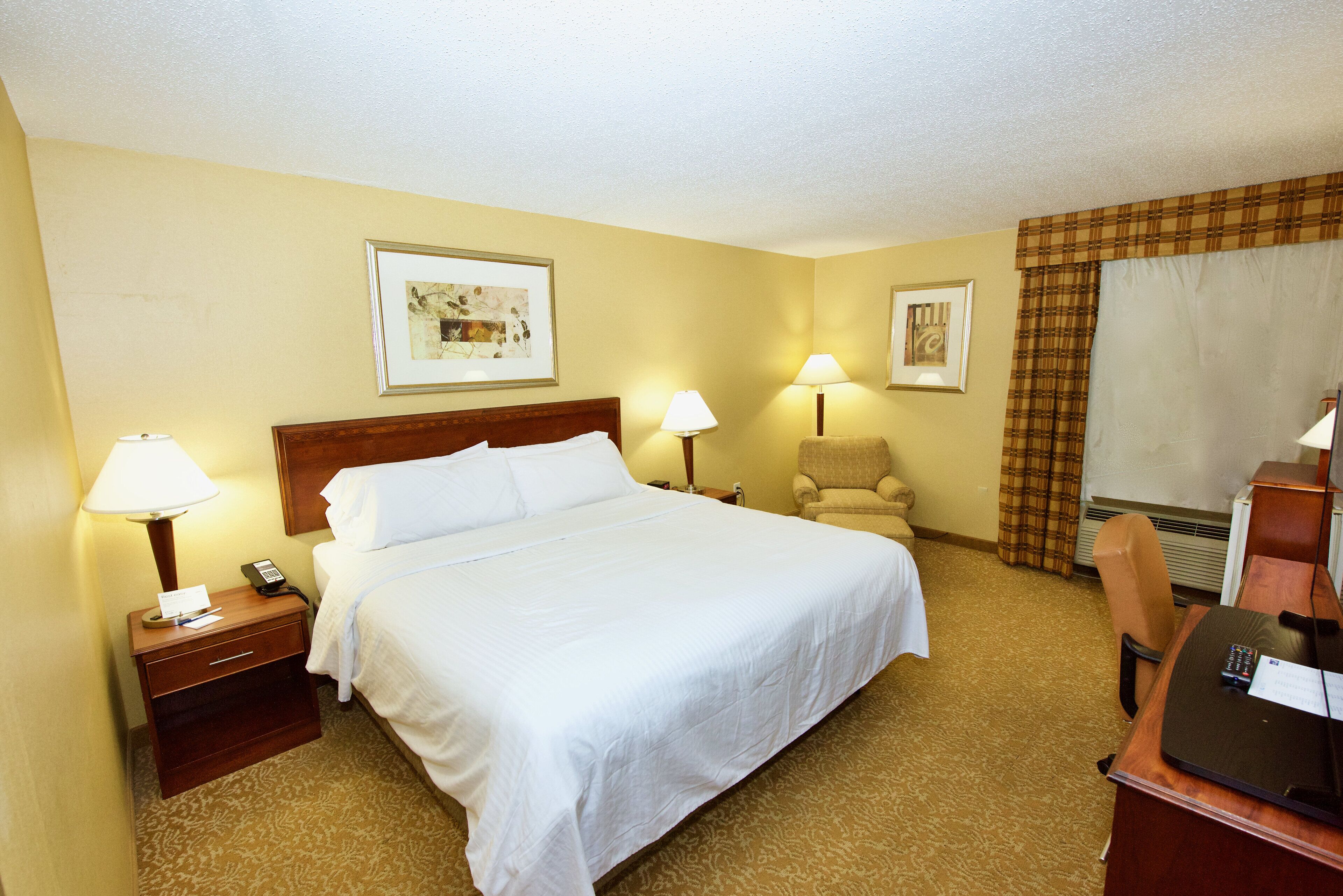 A hotel room with a bed, two bedside lamps, a chair, a desk with a computer, and two framed pictures on the wall.