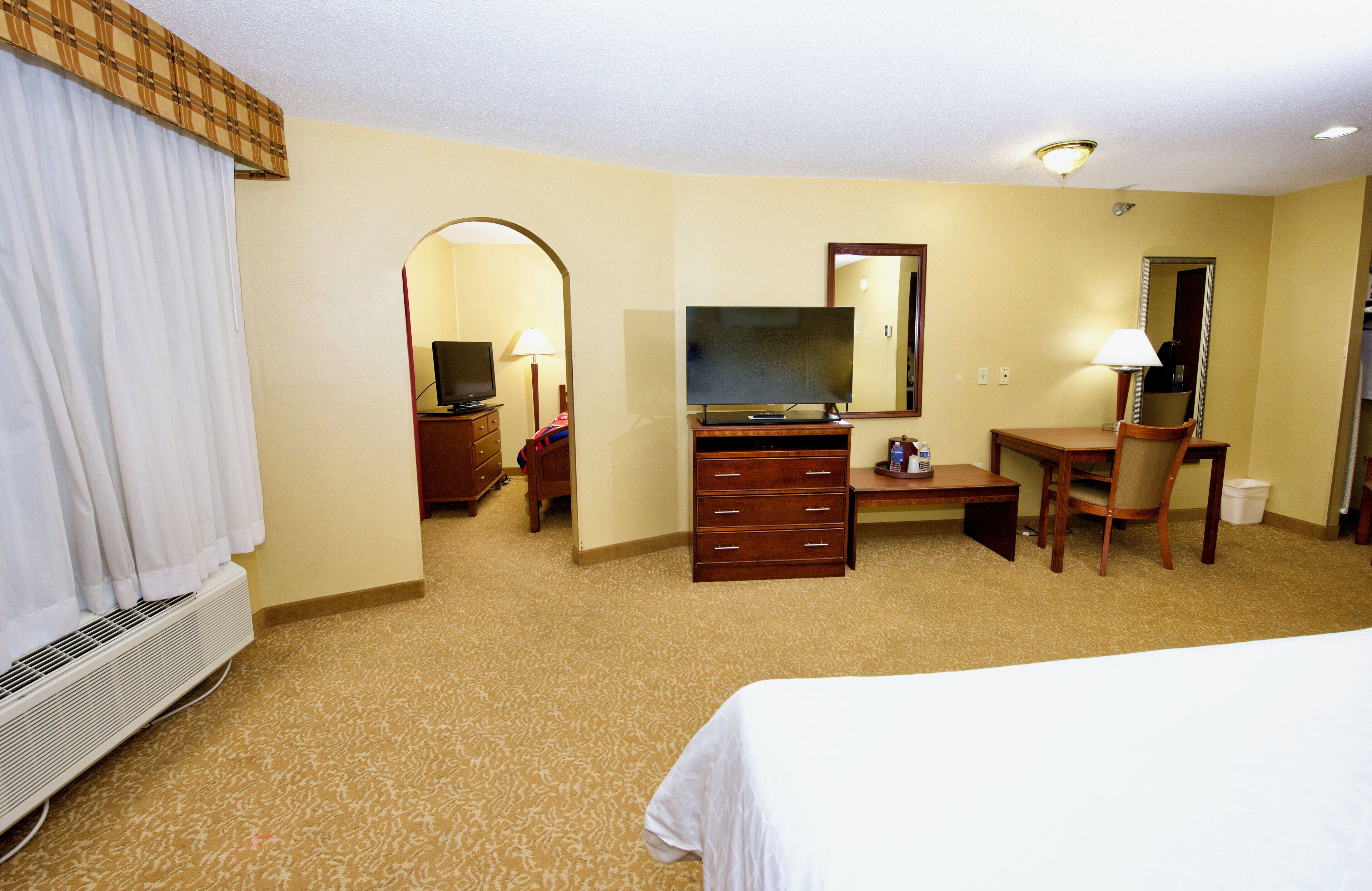 Habitación de hotel con cama, escritorio, computadora, televisor, silla, cómoda y espejo.