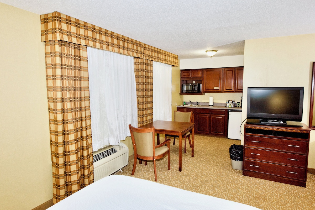 A hotel room with a bed, a dining table with chairs, a kitchen area with a microwave, and a television on a dresser.