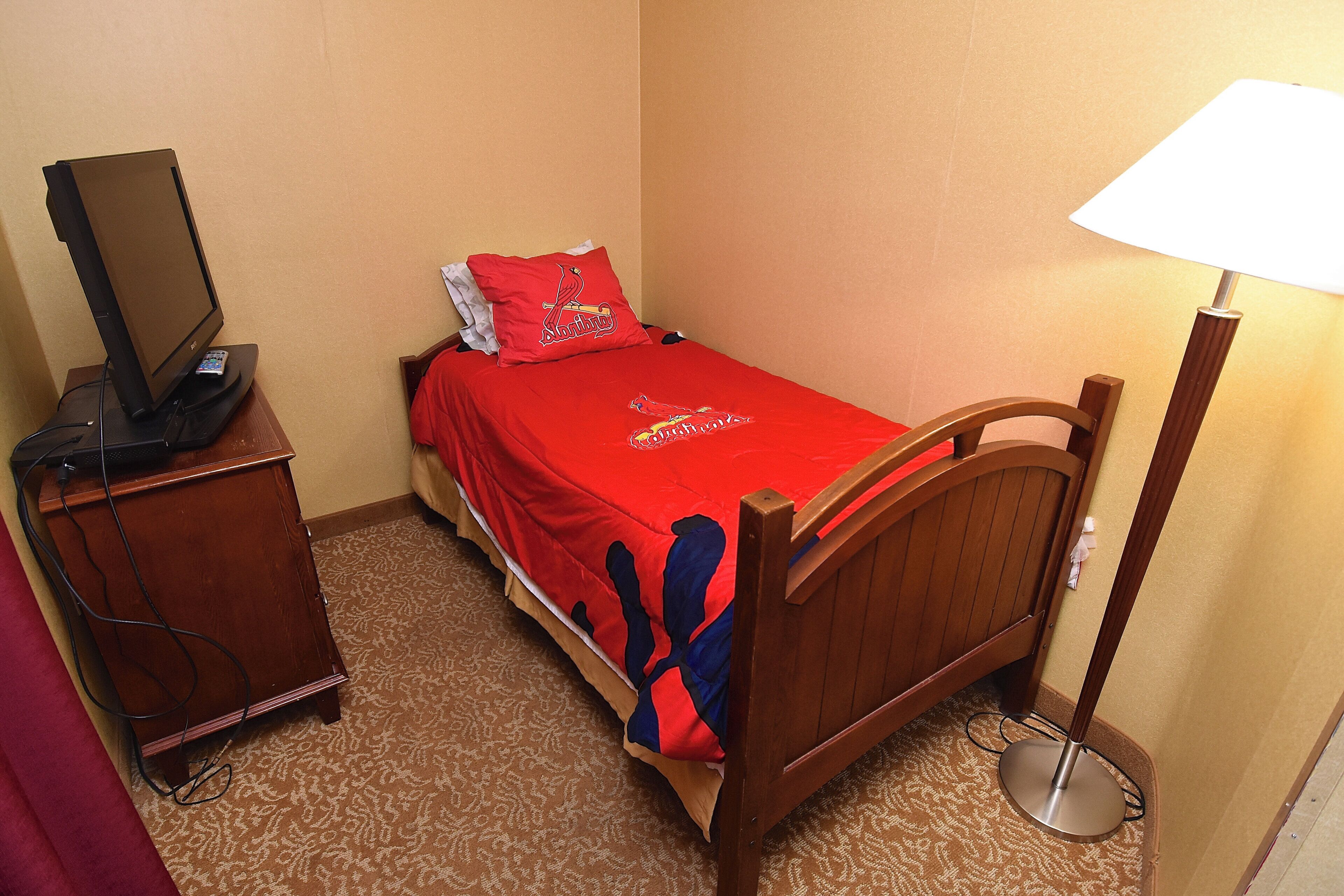 A small bedroom with a single bed, a bedside table, a lamp, and a TV on a stand.