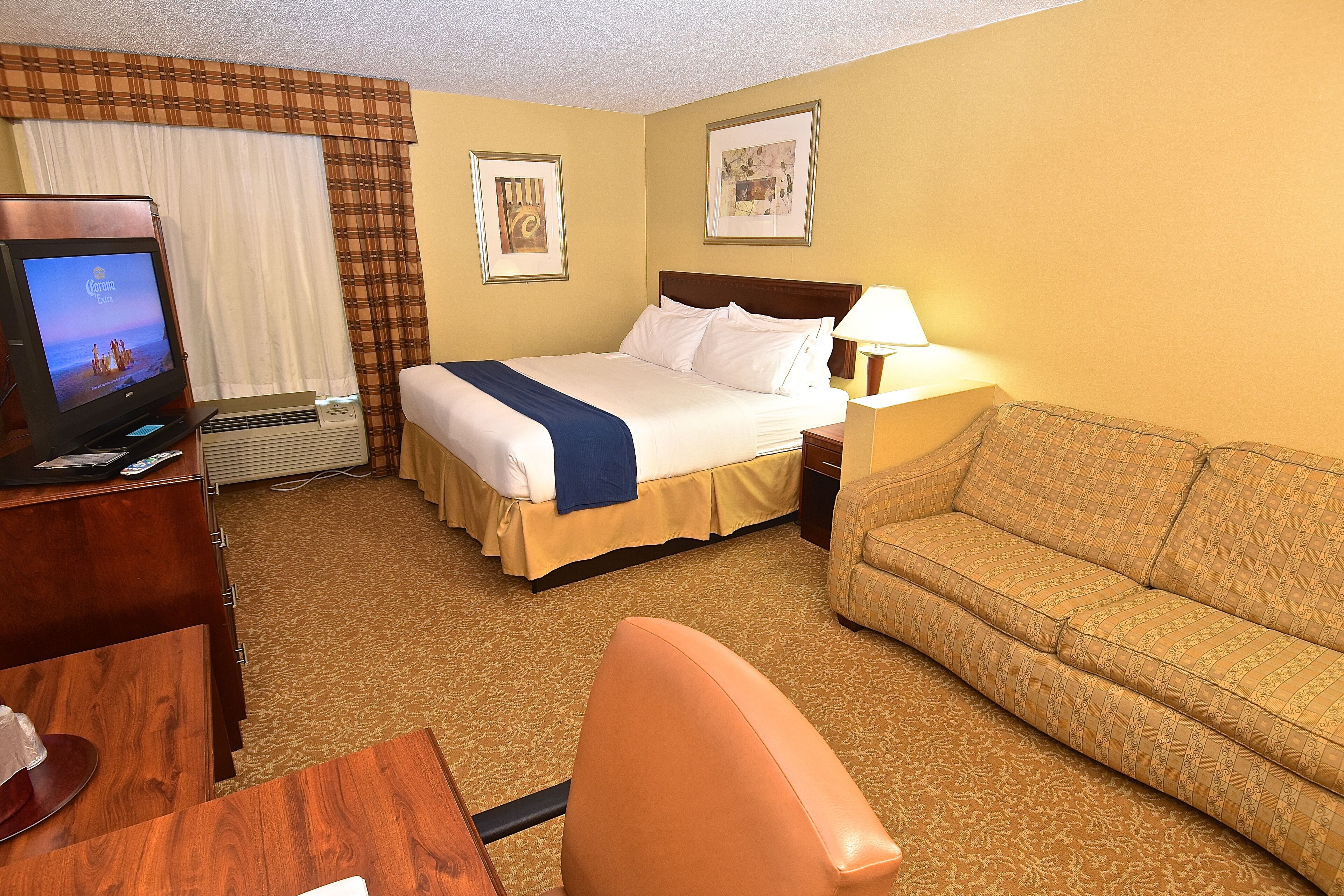 A hotel room with a bed, a sofa, a desk with a computer, and a TV displaying a beach scene.