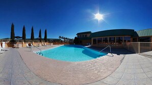 Une piscine baignée de soleil, un espace de repos abrité et de grands arbres en arrière-plan.