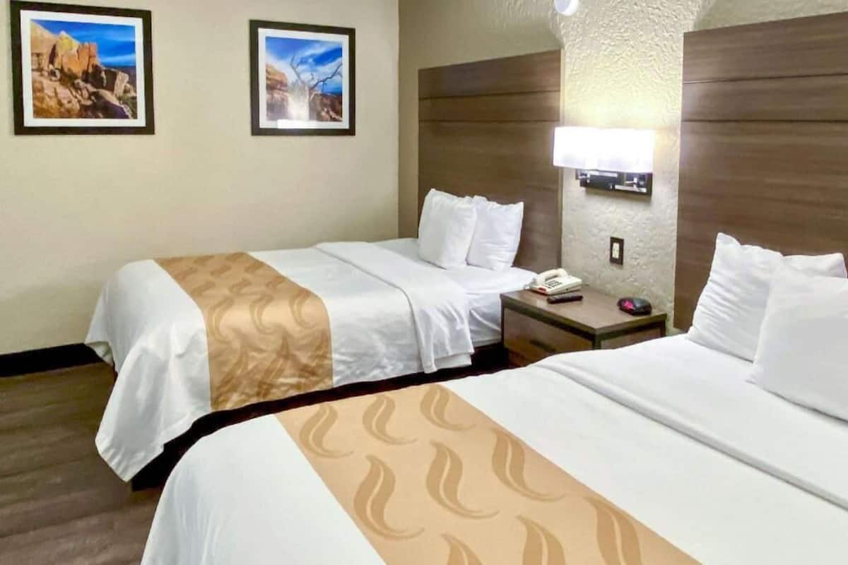 A hotel room with two beds, a nightstand with a phone and a lamp, and two framed pictures on the wall.