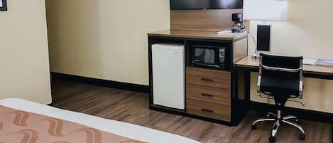 A hotel room with a bed, a desk with a computer, a microwave, a refrigerator, and a TV.