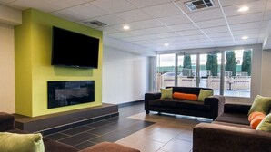 Un hall d’entrée moderne avec un téléviseur à écran plat fixé sur un mur vert, un ensemble de canapés avec des coussins orange et de grandes fenêtres offrant une vue sur l’extérieur.