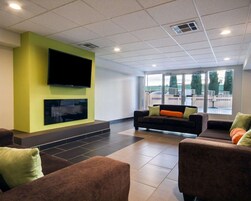 Um lobby moderno com uma TV de tela plana fixada em uma parede verde, um conjunto de sofás com almofadas laranja e grandes janelas com vista para o exterior.
