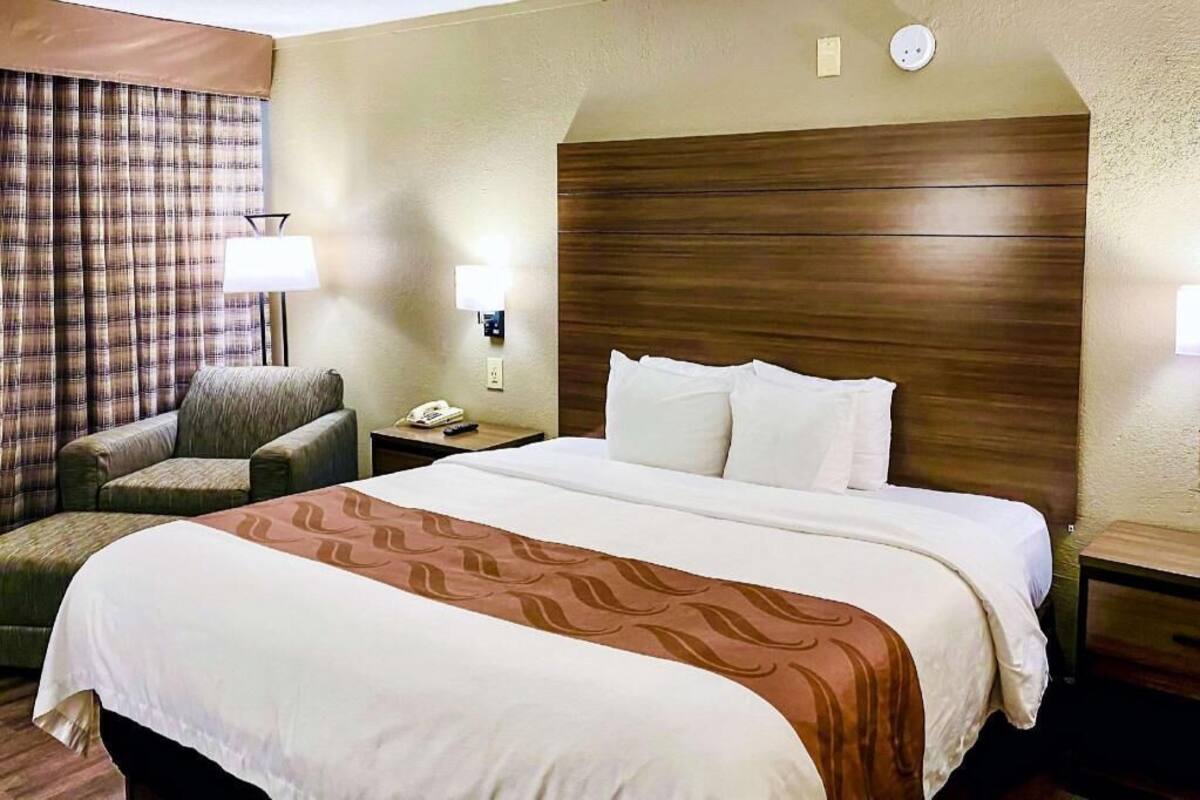 A hotel room with a bed, a chair, a nightstand, a lamp, and a wall-mounted phone.