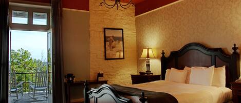 A hotel room with a large bed, a chandelier, a painting on the wall, and a balcony with chairs.