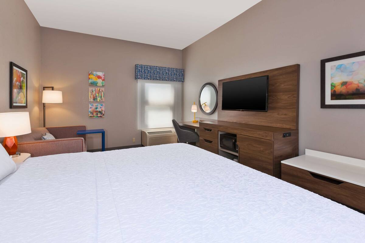 A hotel room with a large bed, a desk with a chair, a flat-screen TV, a desk lamp, and artwork on the walls.
