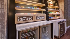A modern kitchenette with built-in appliances, wooden shelves with snacks, and a refrigerator stocked with beverages. - Hampton Inn Princeton (Princeton)