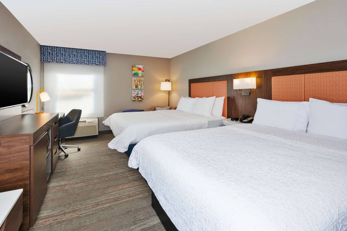 A hotel room with two beds, a desk, a chair, a TV, and artwork on the wall.
