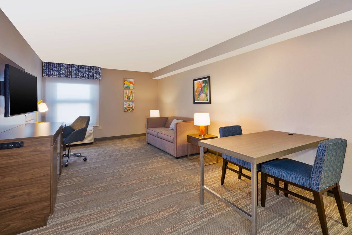 A hotel room with a wooden desk, a sofa, a dining table with chairs, a TV, and artwork on the walls.