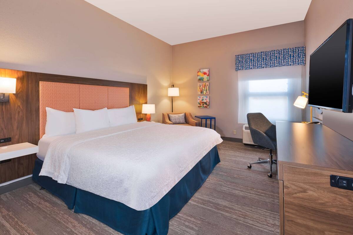 A hotel room with a large bed, bedside tables, a desk, a chair, a TV, and a window with a view.