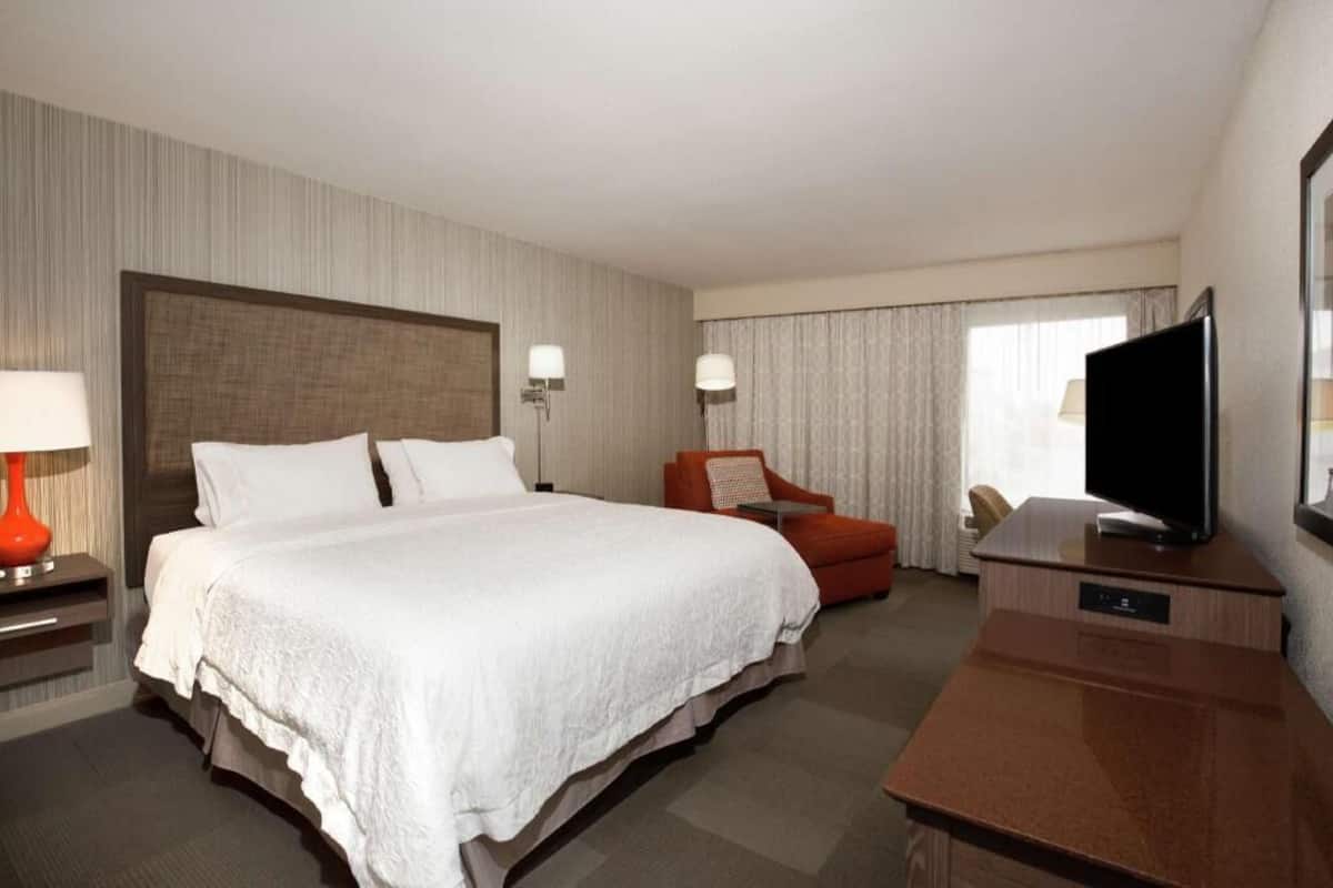 A hotel room with a large bed, a desk with a flat-screen TV, a chair, and a lamp.